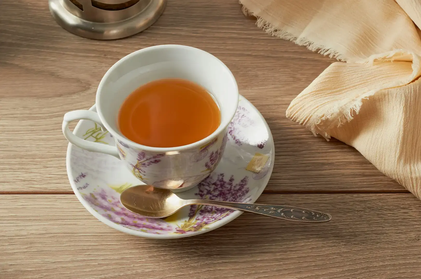 A white porcelain teacup with a delicate purple lavender floral pattern filled with clear golden-amber tea, resting on a matching saucer alongside a small silver spoon on a light wooden surface next to a soft cream-colored cloth.