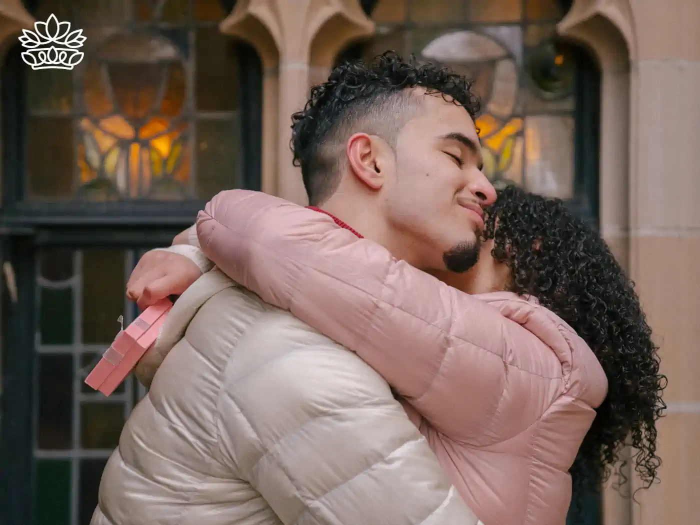 A young man with curly hair hugging a woman with curly hair, holding a pink gift box behind her back, expressing affection and joy. A stained glass window forms the backdrop. 