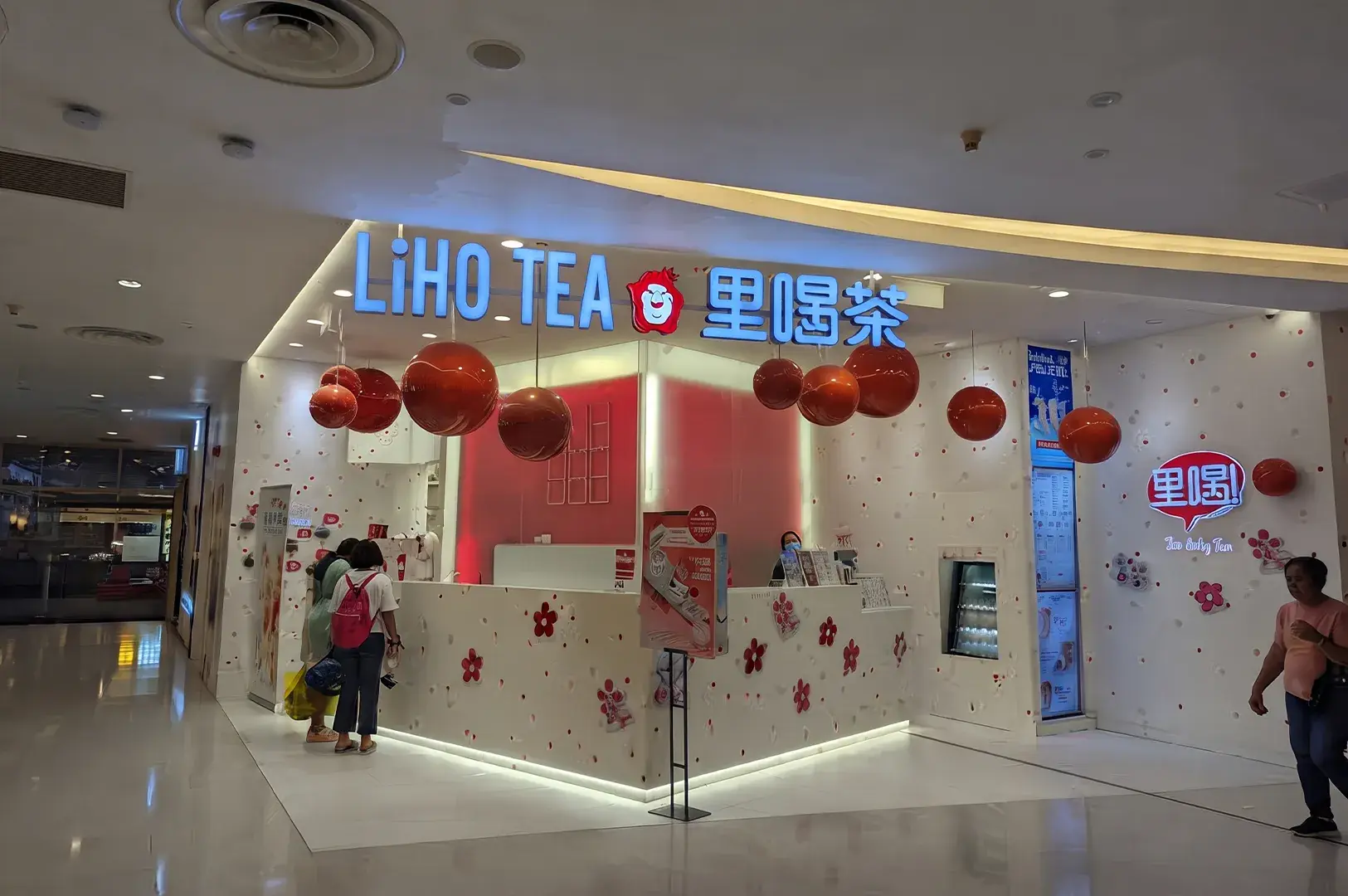 Storefront of a LiHO TEA outlet inside a shopping mall, featuring bright illuminated signage, suspended red spherical decorations, and a white service counter decorated with floral accents.