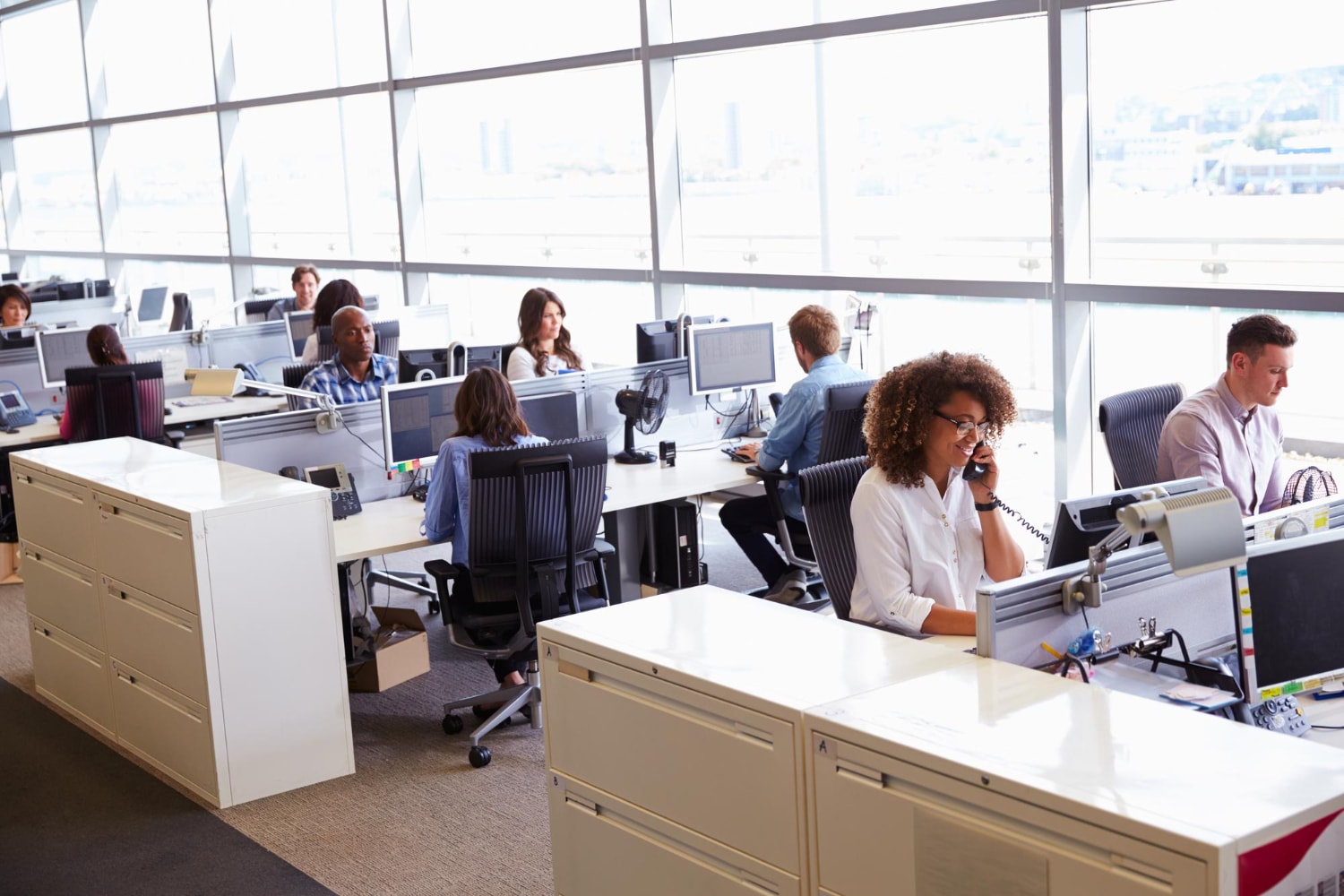 A spacious office with multiple employees working at their desks, engaging in phone calls and computer-based tasks. The open-plan design suggests a Cloud communications infrastructure supported by Business VoIP providers.