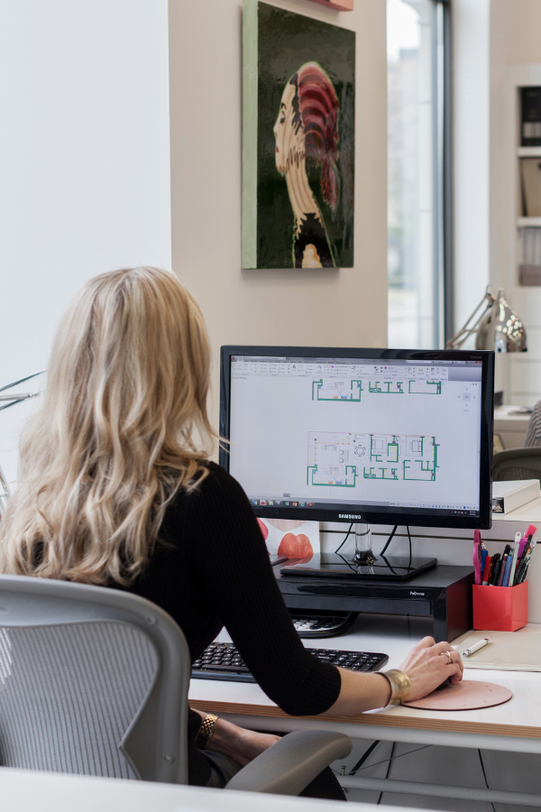 A woman works at a computer displaying architectural floor plans in a bright design studio, with art and office supplies around her desk.