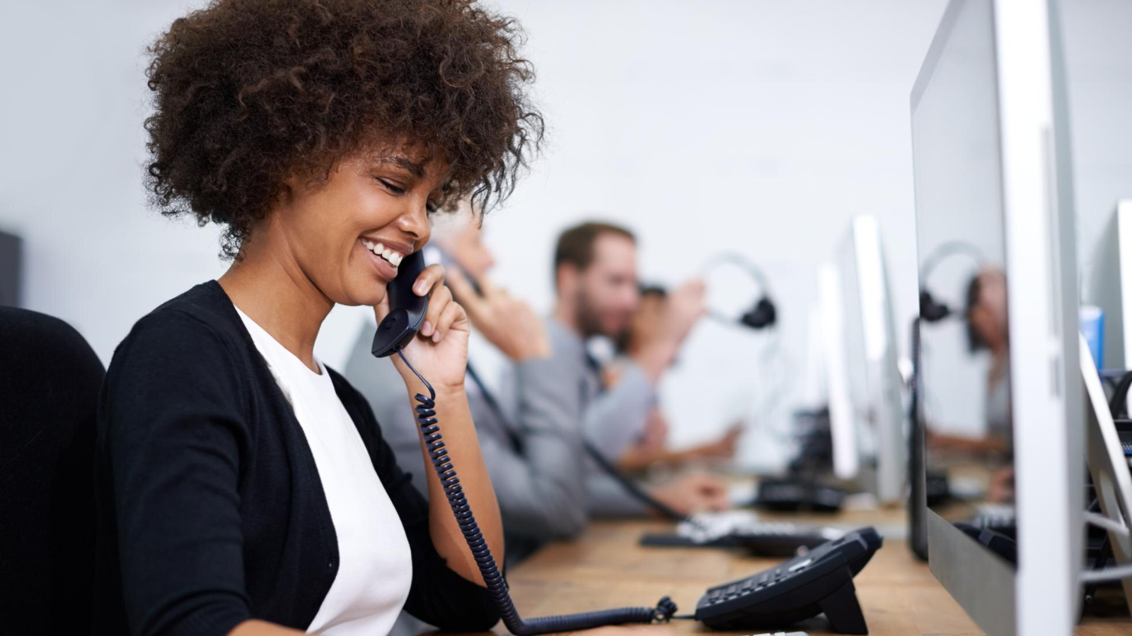 A smiling woman with curly hair speaks on a desk phone in a busy, modern office environment. She is surrounded by coworkers on similar calls, depicting the practical use of VoIP and cloud-based communication for streamlined, cost-effective telecom operations.