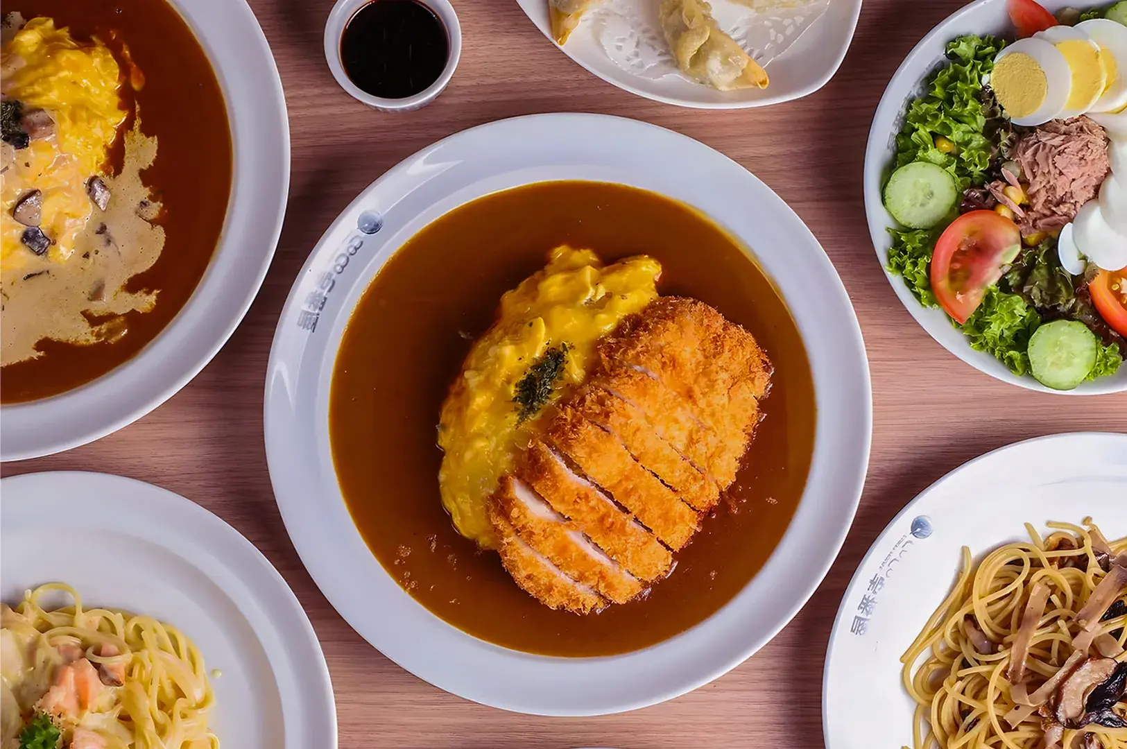 A table with various dishes including a centerpiece of crispy fried cutlet on curry rice with omelet. Surrounding plates have pasta, salad, and dumplings.