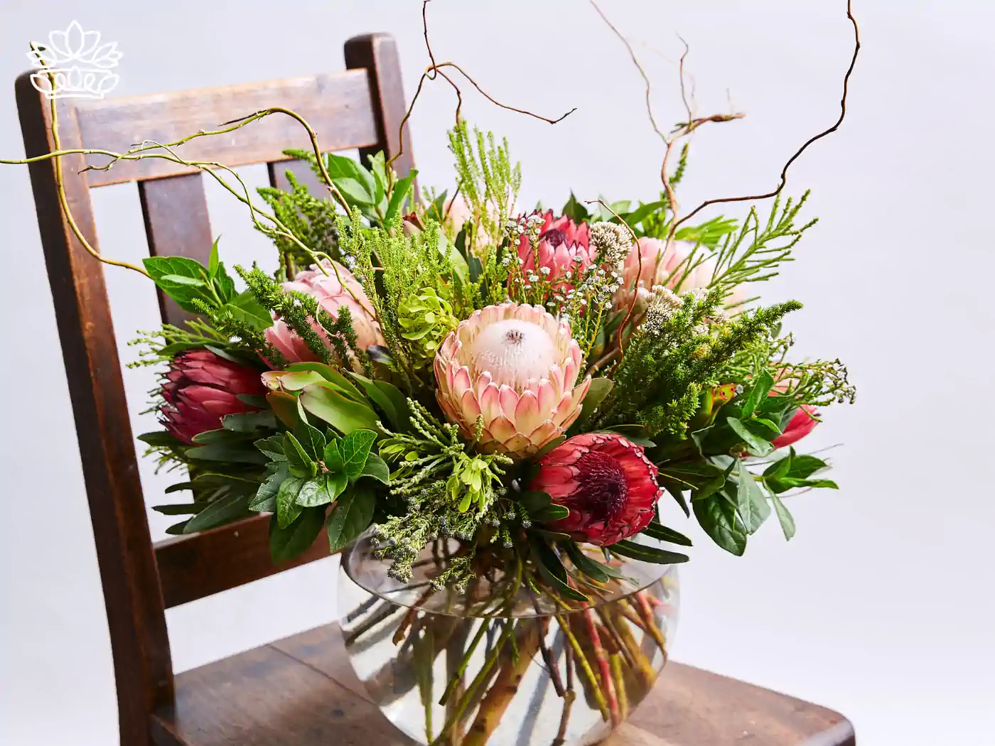 Exquisite arrangement of proteas and mixed foliage in a glass vase, artistically placed on a wooden chair. Fabulous Flowers and Gifts, Guest House and Hotel Gift Boxes Delivered with Heart.