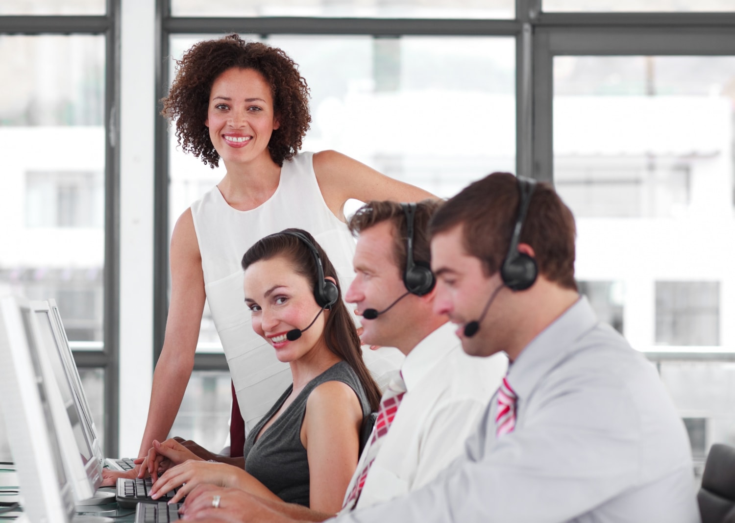 A group of professionals in a bright, open-plan office, each wearing headsets and working on computers. Their team leader stands behind them, smiling, showcasing real-time contact center analytics in action to improve customer engagement.