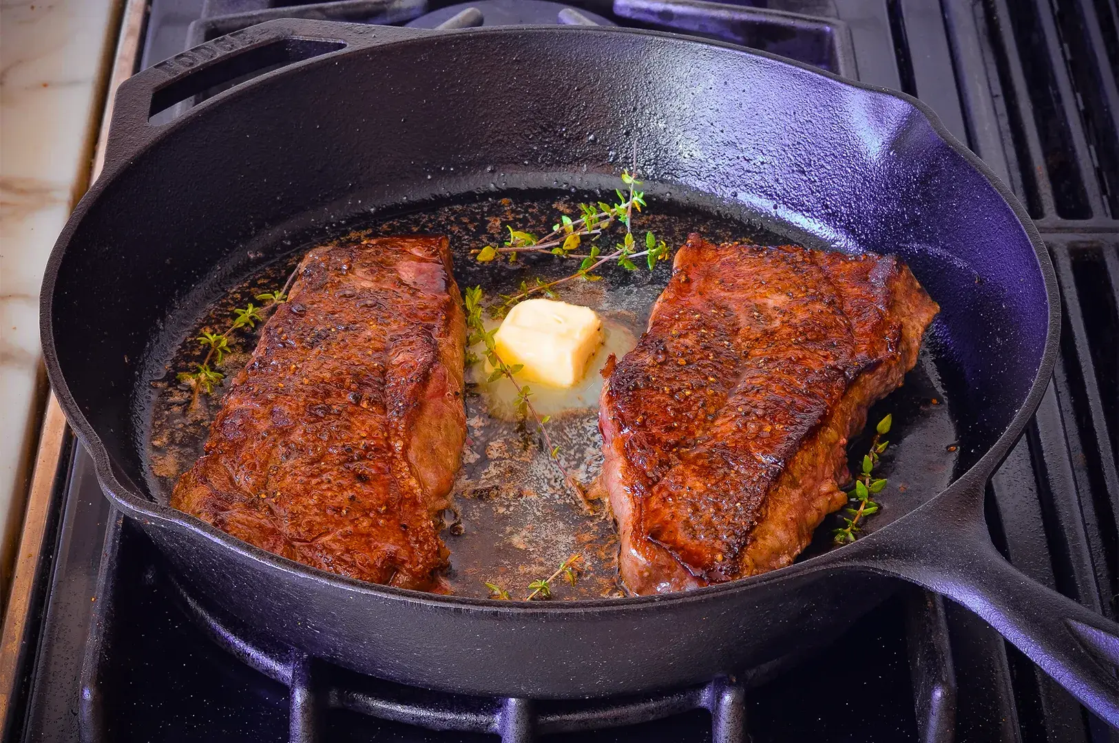 Two juicy steaks are sizzling in a cast iron skillet on a stovetop, accompanied by melting butter and sprigs of fresh thyme, evoking a savory aroma.