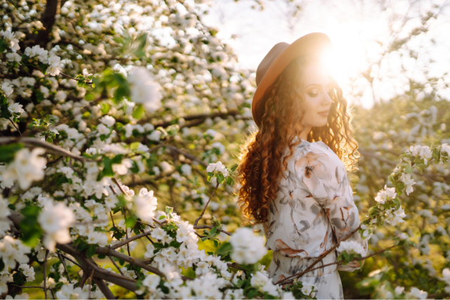 Rothaarige Frau in Frühlingskleidung mit Hut steht zwischen blühenden Baumästen