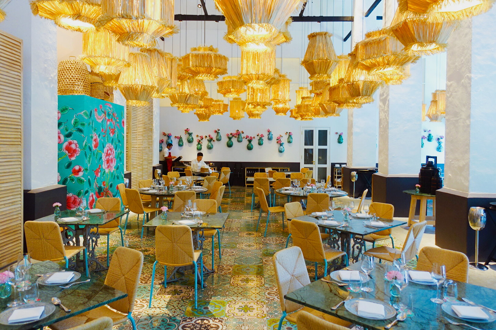 Bright, elegant restaurant with patterned tiles, wicker chandeliers, and vibrant floral wall art. Tables are set for dining, creating a welcoming atmosphere.
