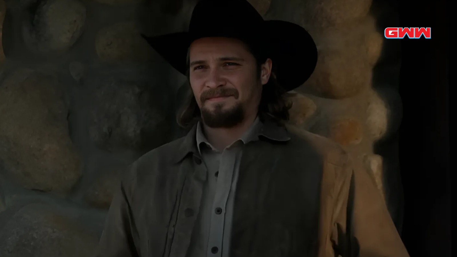 Luke Grimes as Kayce Dutton smiling outside rustic stone cabin