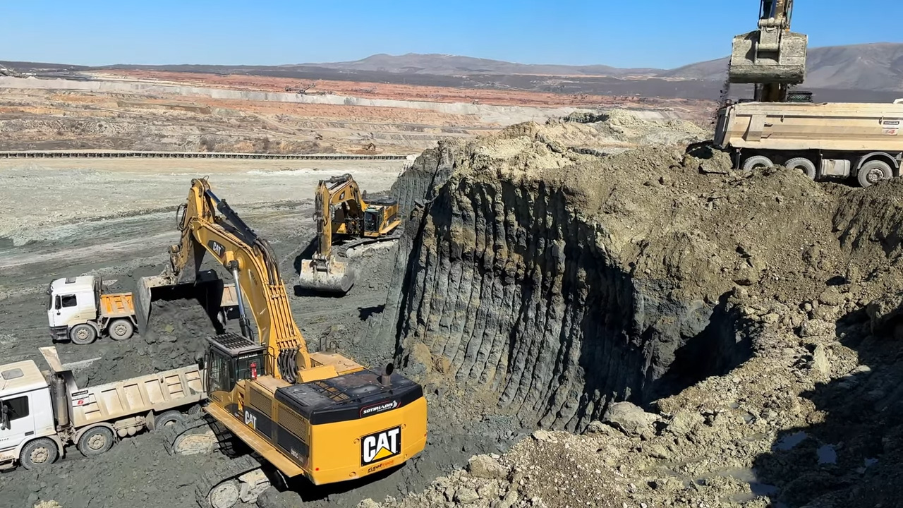 Construction equipment, including excavators and dump trucks, working in a large open-pit mining site with rocky terrain and a clear blue sky