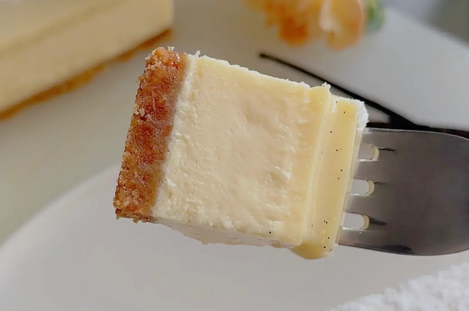 A fork holds a creamy cheesecake bite with a golden crumb crust against a white plate, in warm light that makes the dessert look rich and inviting.