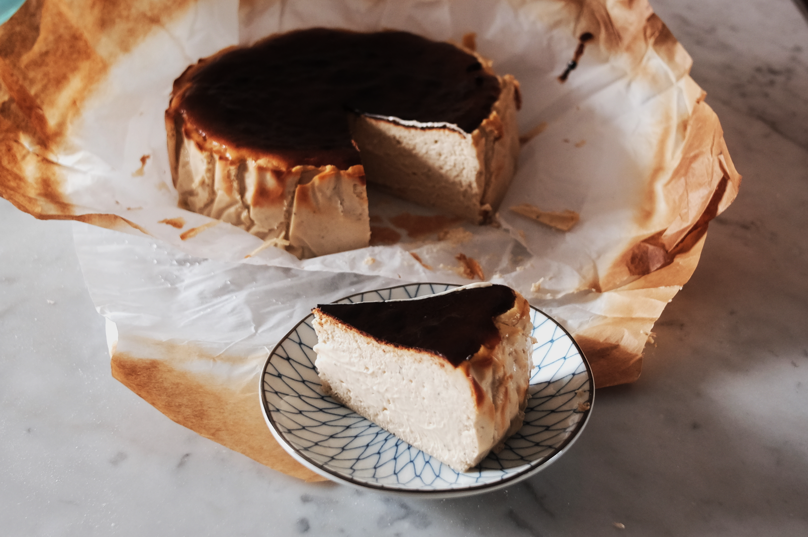 Burnt Basque cheesecake with a dark caramelized top and creamy interior on parchment paper. A slice sits on a patterned plate. Elegant and rustic.