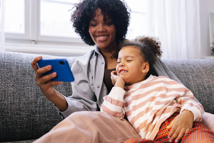 Pretty mom in a gray jacket sitting on the sofa with her little girl and snapping a selfie.