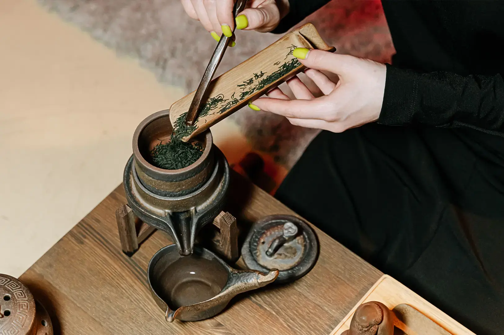 Features a person with bright yellow nail polish using a small wooden tool to carefully scoop dark, dried green tea leaves from a rectangular wooden tray into a traditional, rustic ceramic tea-making vessel on a wooden table.