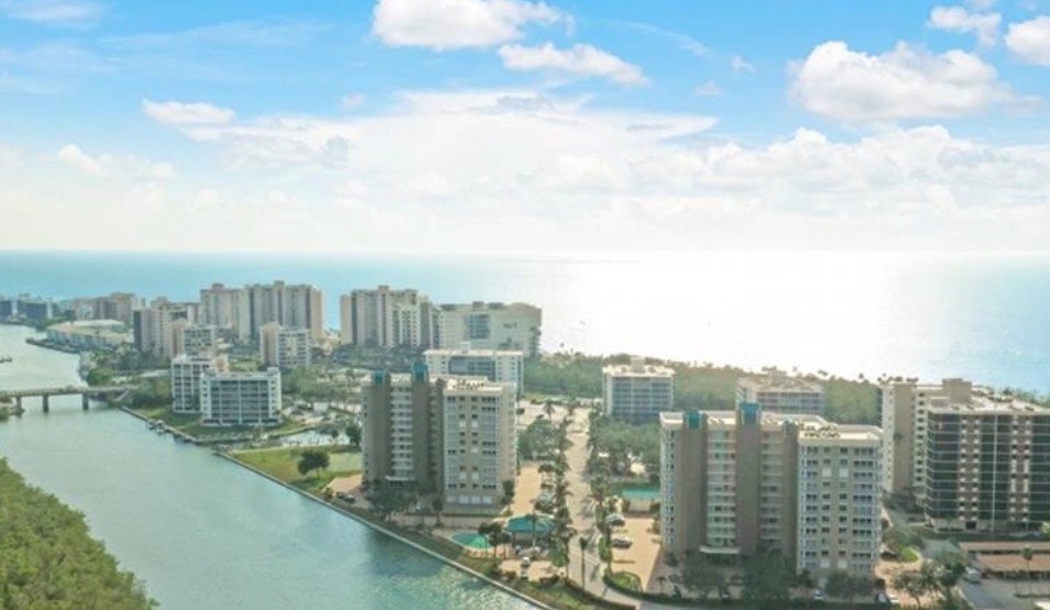 The image showcases a modern condominium interior with floor-to-ceiling windows that allow for abundant natural light, highlighting the contemporary furnishings and spacious floor plan. This exquisite turnkey beachfront condominium, located in the Vanderbilt Beach neighborhood of Naples, FL, offers breathtaking panoramic views and a luxurious lifestyle.