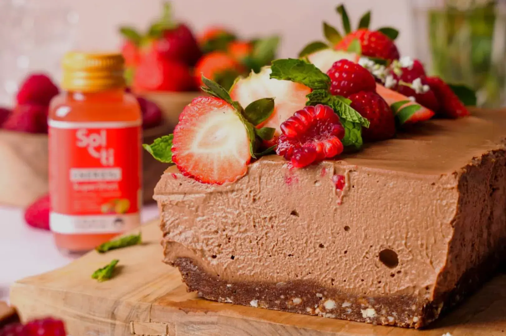 A delicious chocolate mousse cake topped with strawberries, raspberries, and mint leaves, with a bottle of drink in the background.