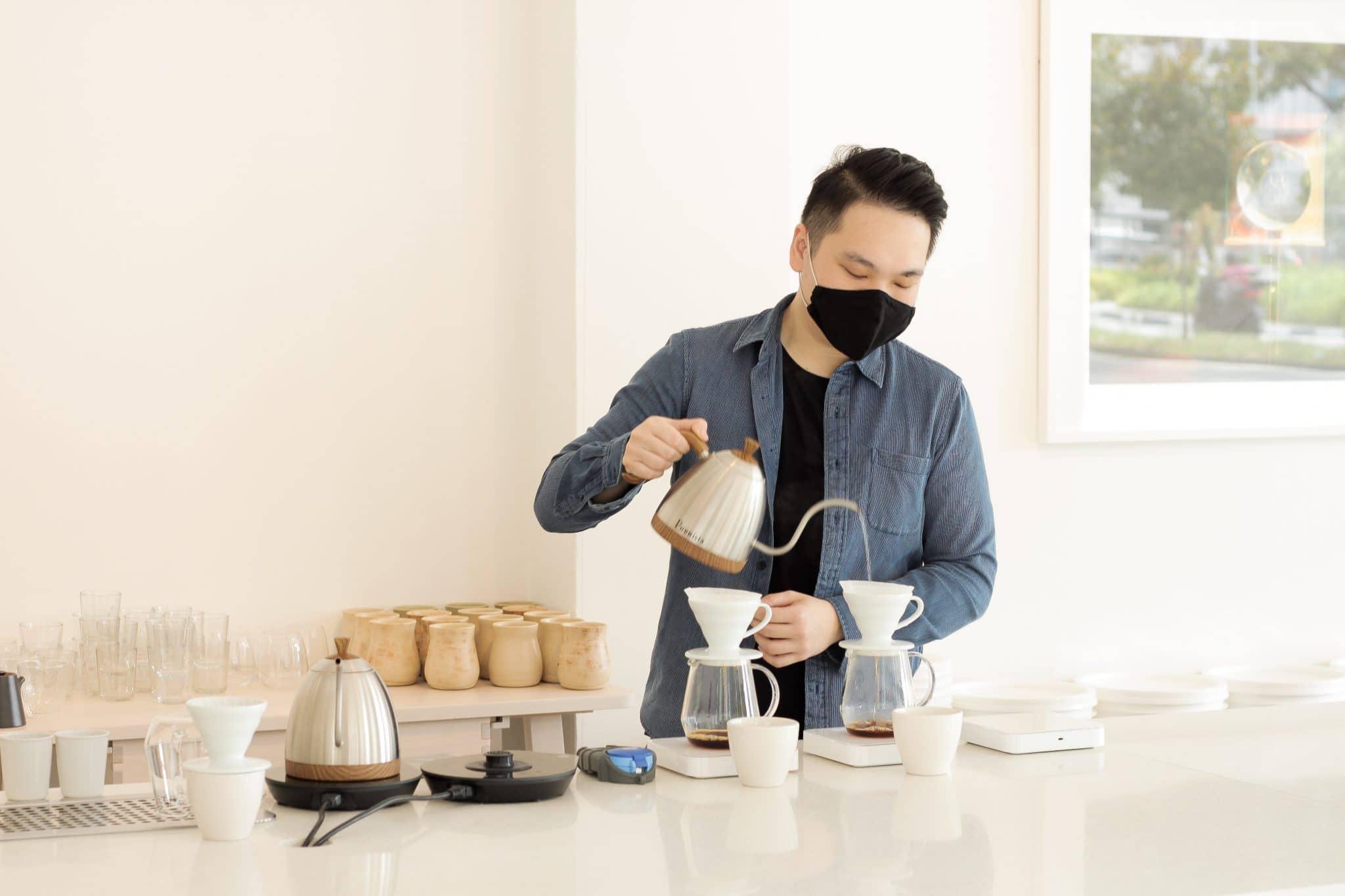 A barista in a blue corduroy shirt and black face mask carefully pours water from a gooseneck kettle into two pour-over coffee drippers. The scene is set in a bright, minimalist cafe featuring white surfaces and organized rows of ceramic cups and glassware.