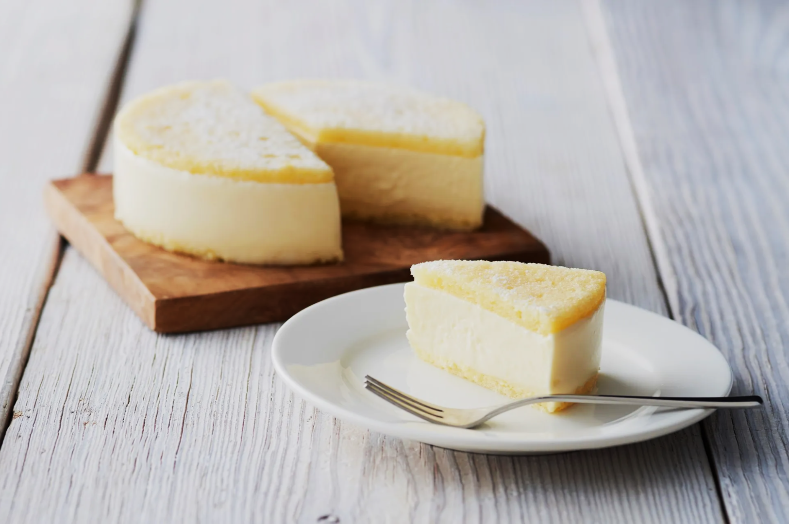 A creamy cheesecake slice rests on a white plate with a fork, set on a rustic white wooden table. The remaining cake is on a wooden board behind.