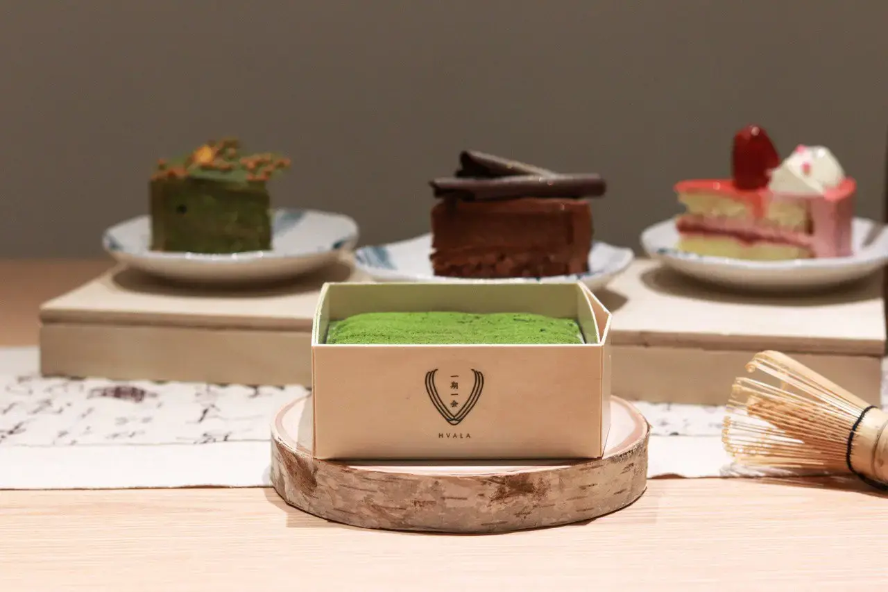 In the image, a vibrant green matcha tiramisu is featured in a small wooden box labeled with the "Hvala" logo, resting on a decorative birch wood slice. In the blurred background, three other assorted cakes—green tea, chocolate, and strawberry—are displayed on small plates atop a wooden riser.