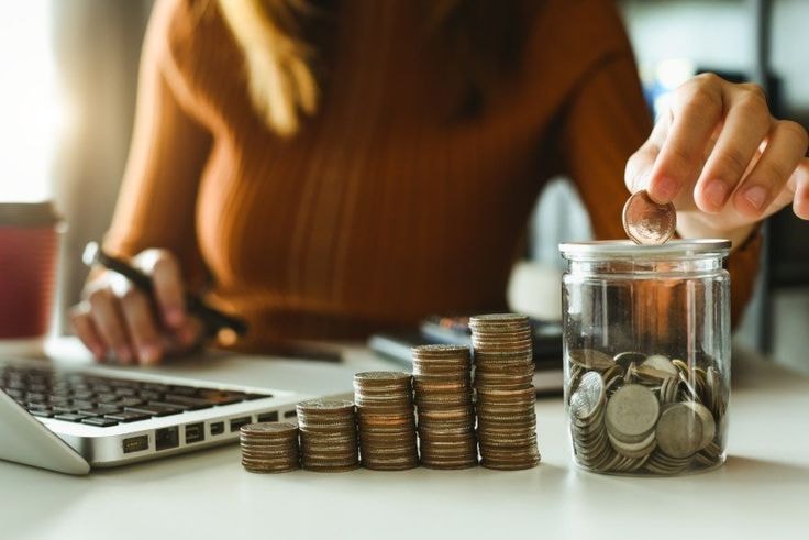 Kobieta siedzi przy laptopie i robi podsumowanie finansowe. Wrzuca monetę do słoika z innymi drobniakami.