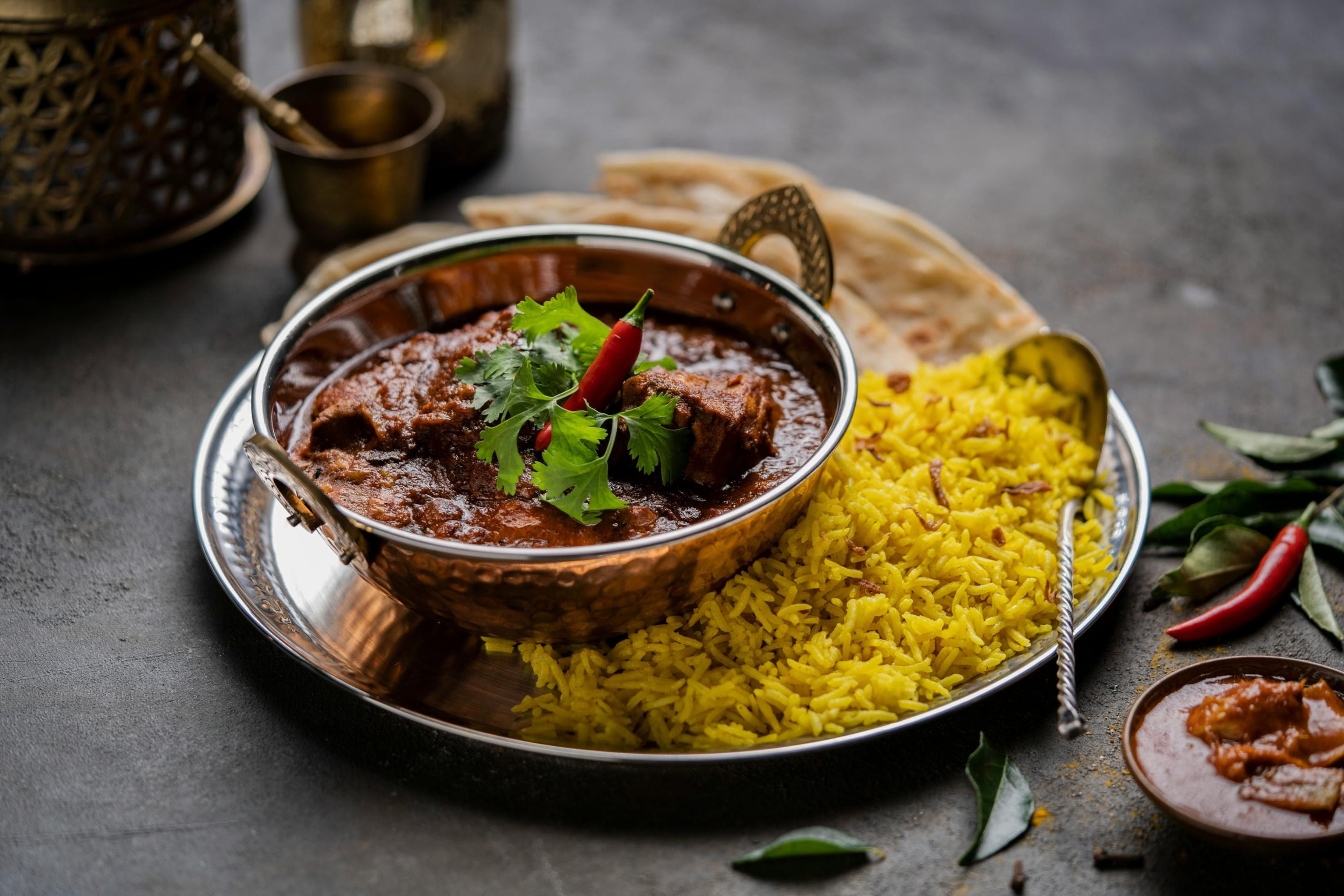 This image features a copper bowl filled with a rich, dark meat curry topped with a fresh red chili and cilantro, served alongside a mound of vibrant yellow saffron rice on a silver platter. Accompanied by a piece of flatbread and a small side dish, the meal is presented on a dark, textured surface with decorative brass elements in the background.