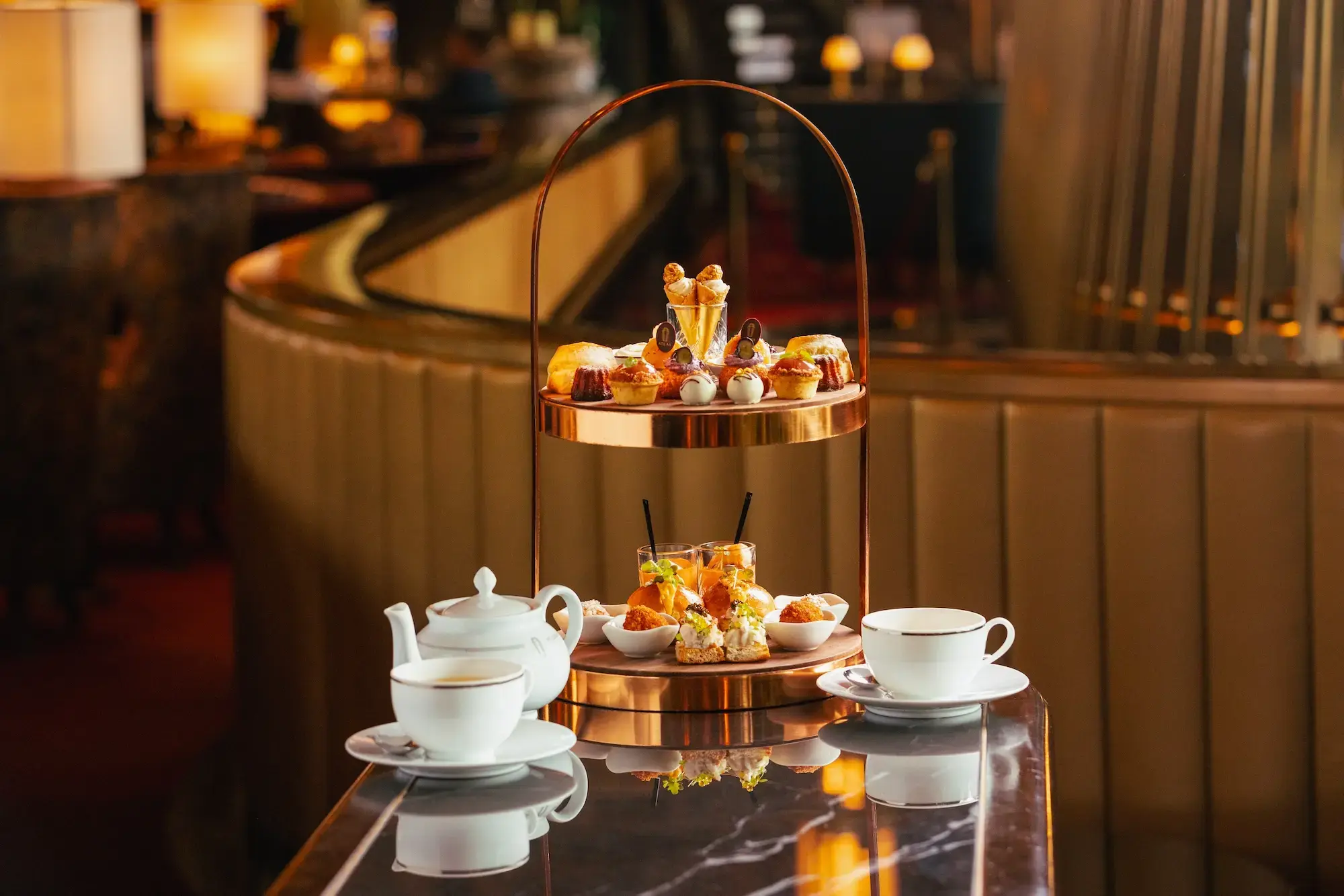 An elegant two-tiered afternoon tea stand is presented on a reflective marble table, showcasing a variety of sweet pastries on top and savory bites below. A white teapot and two matching teacups on saucers flank the stand, all set against a warm, softly blurred Art Deco restaurant interior.