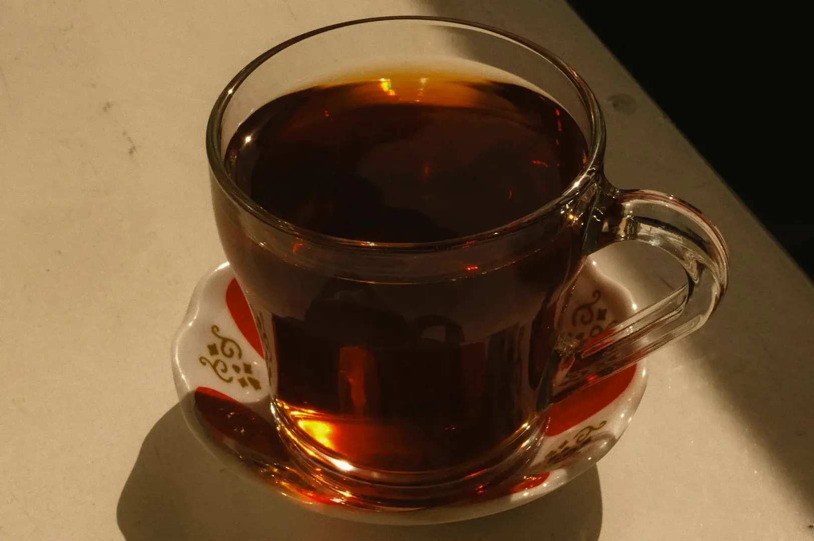 A close-up, side-angle shot displays a clear glass teacup filled to the brim with dark amber-colored tea, sitting on a small, white ceramic saucer adorned with delicate red and gold abstract leaf patterns. The cup features a sturdy, curved glass handle on the right and rests on a light-colored, slightly reflective countertop, bathed in warm, directional lighting from the lower left that catches the base of the glass in a bright orange glow and casts a soft shadow to the left.