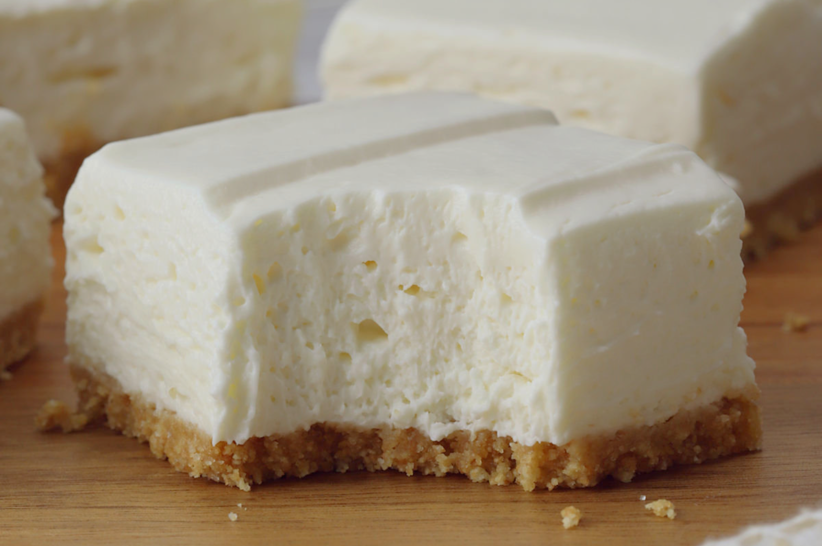A creamy slice of cheesecake with a crumbly graham cracker crust on a wooden surface. The texture looks smooth and rich, conveying a sense of indulgence.