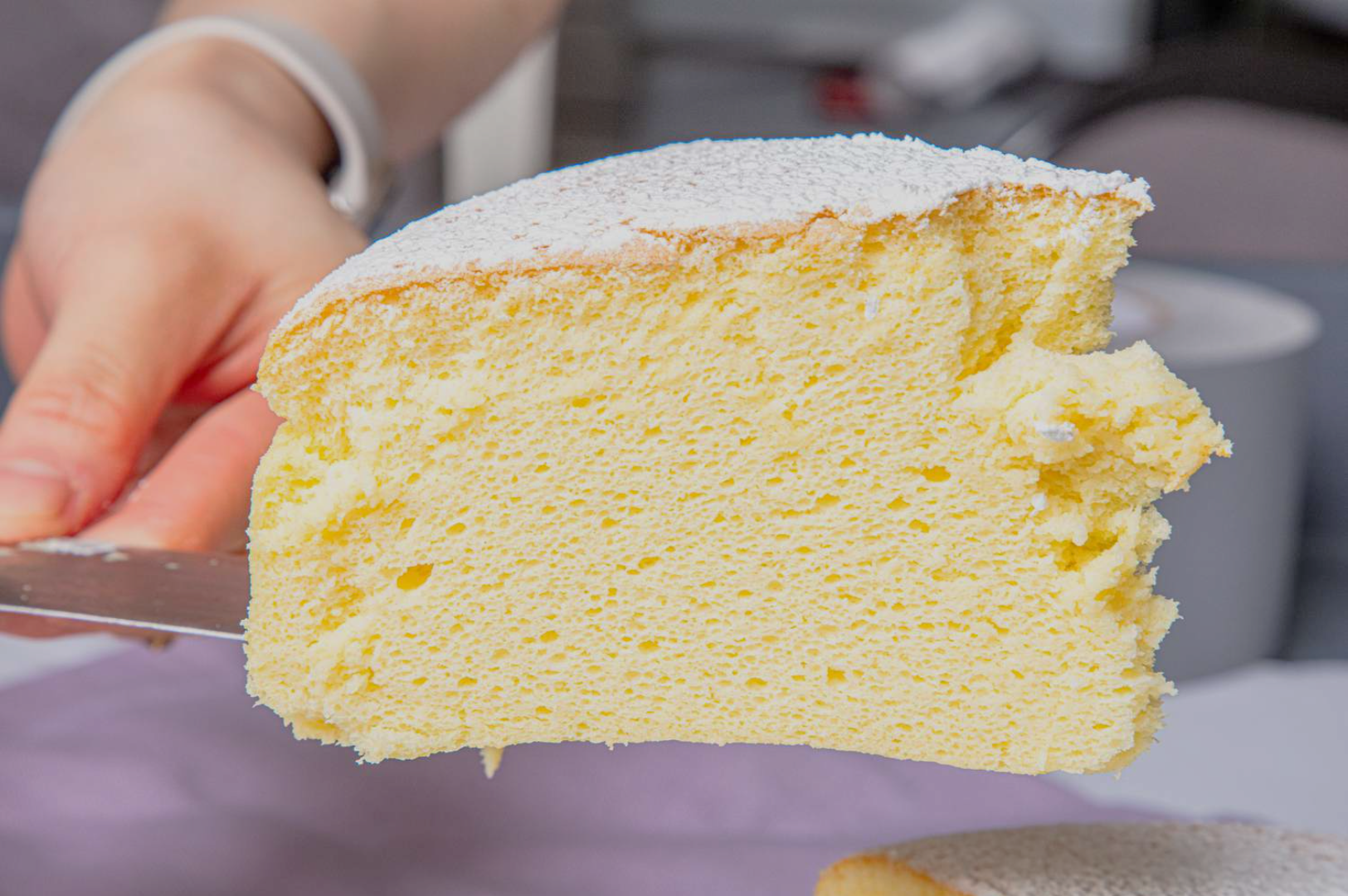 A hand holds a slice of fluffy, yellow sponge cake dusted with powdered sugar. The cake looks soft and airy, evoking a sense of lightness and sweetness.