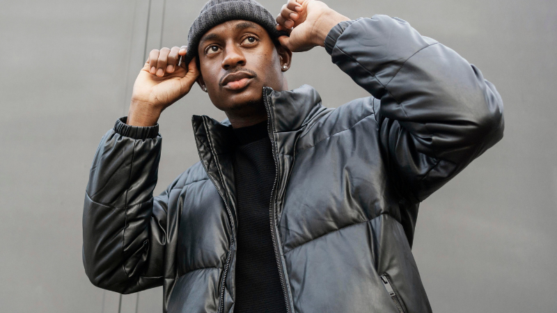 A person wearing a black puffer gorpcore jacket and a beanie, adjusting their hat with both hands.