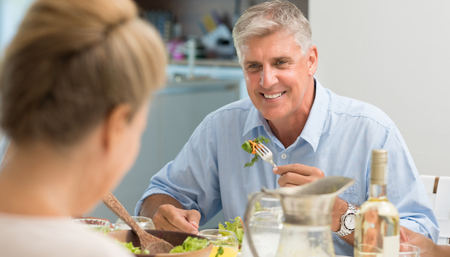 Smilende eldre mann spiser salat ved middagsbordet sammen med en kvinne, omgitt av juice, vin og frisk mat i lyst kjøkken. Ask ChatGPT