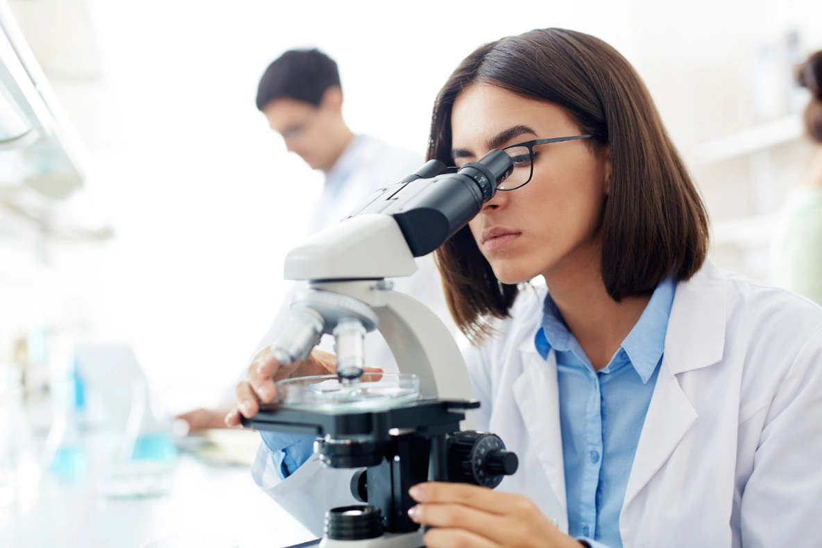 A person in a lab coat looks into a microscope.