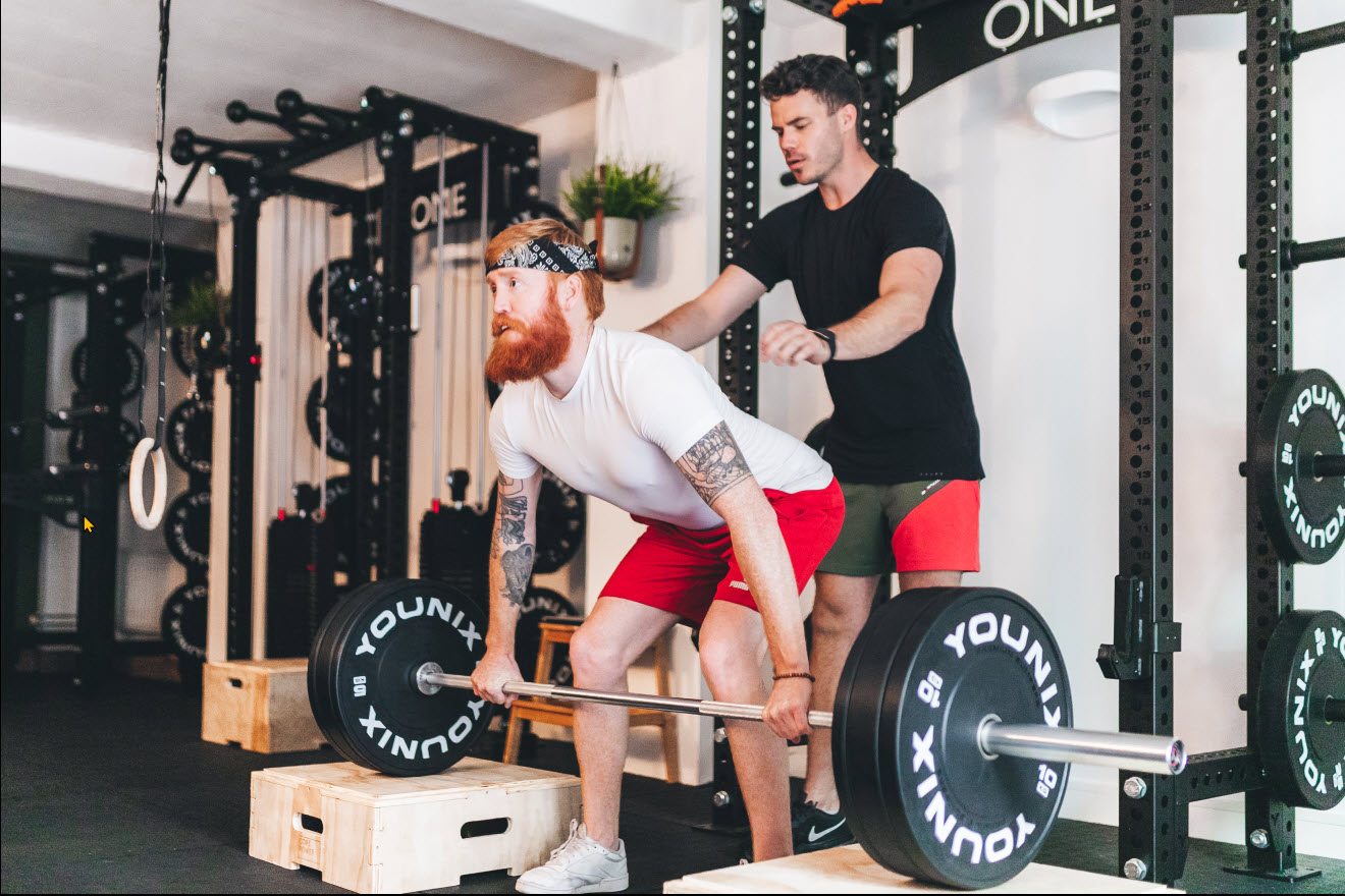 "A male personal trainer guiding a male client through a deadlift exercise in a gym setting"