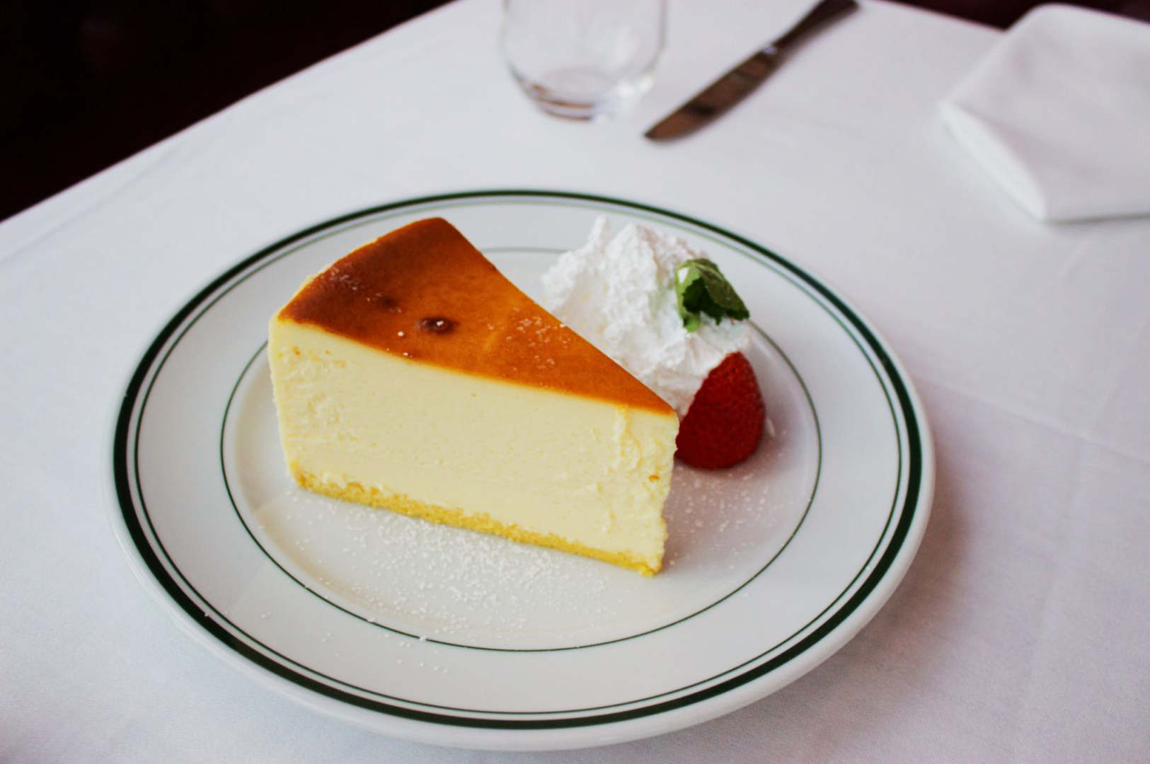 A slice of creamy cheesecake sits on a white plate with green trim, topped with whipped cream and a fresh strawberry. The setting is elegant with a white tablecloth.