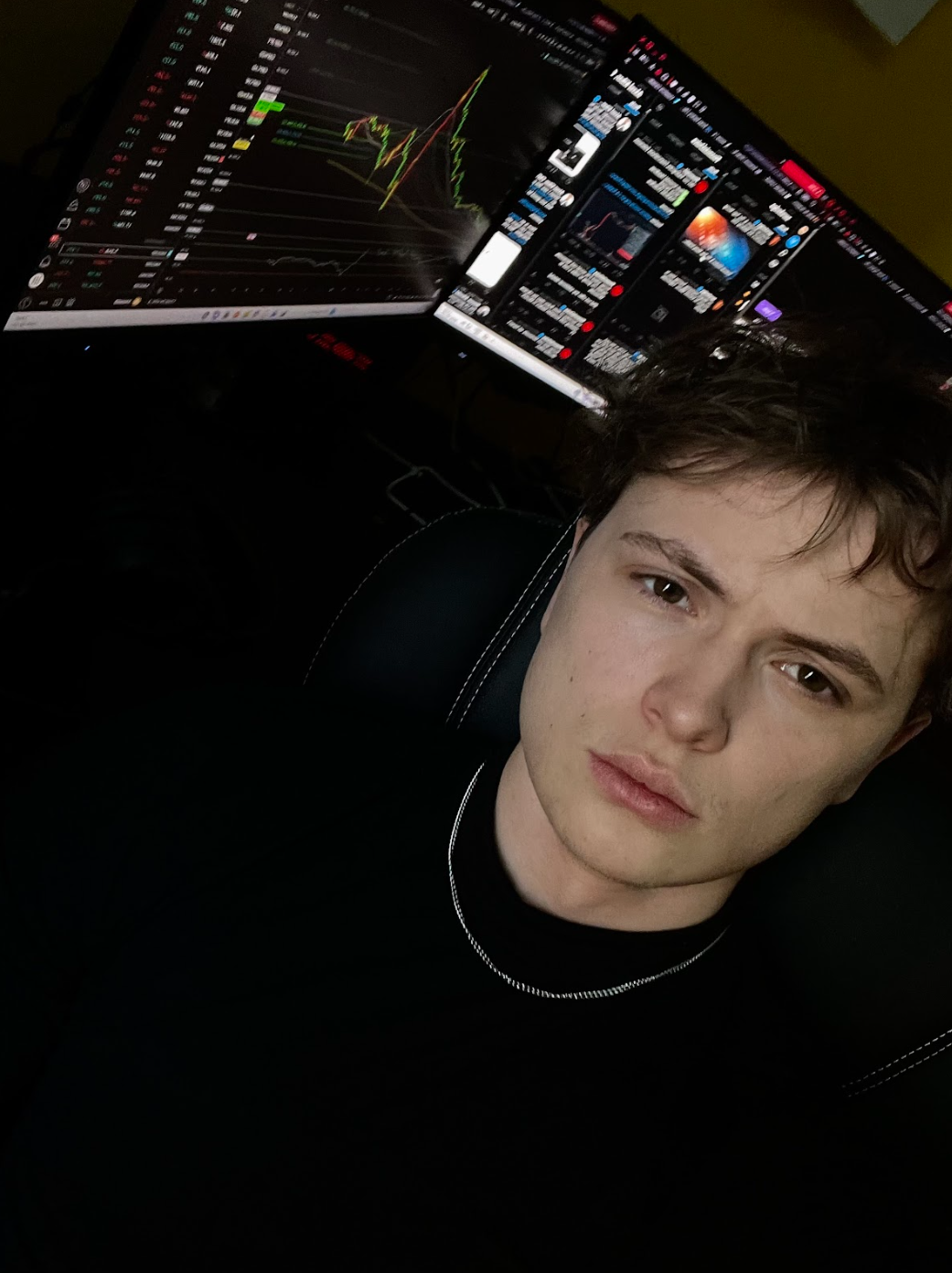 Young male trader seated in a gaming chair at a dimly lit desk, gazing directly into the camera with a calm, focused expression, while multiple monitors behind him display live trading charts and market data — evoking the solitary, late-night grind of an independent retail trader.