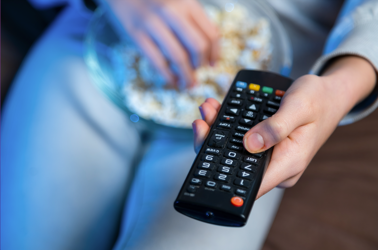 one hand holds a tv remote while another hand grabs popcorn