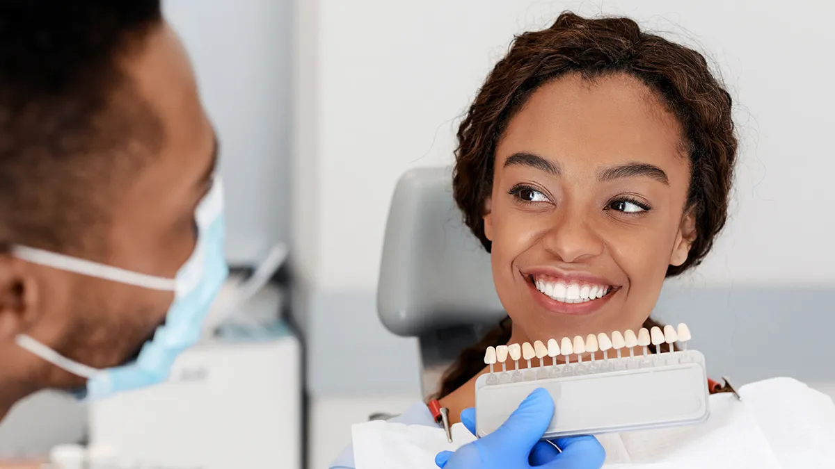 A patient with a bright smile looking at a dentist who is holding a tooth shade guide for color matching.
