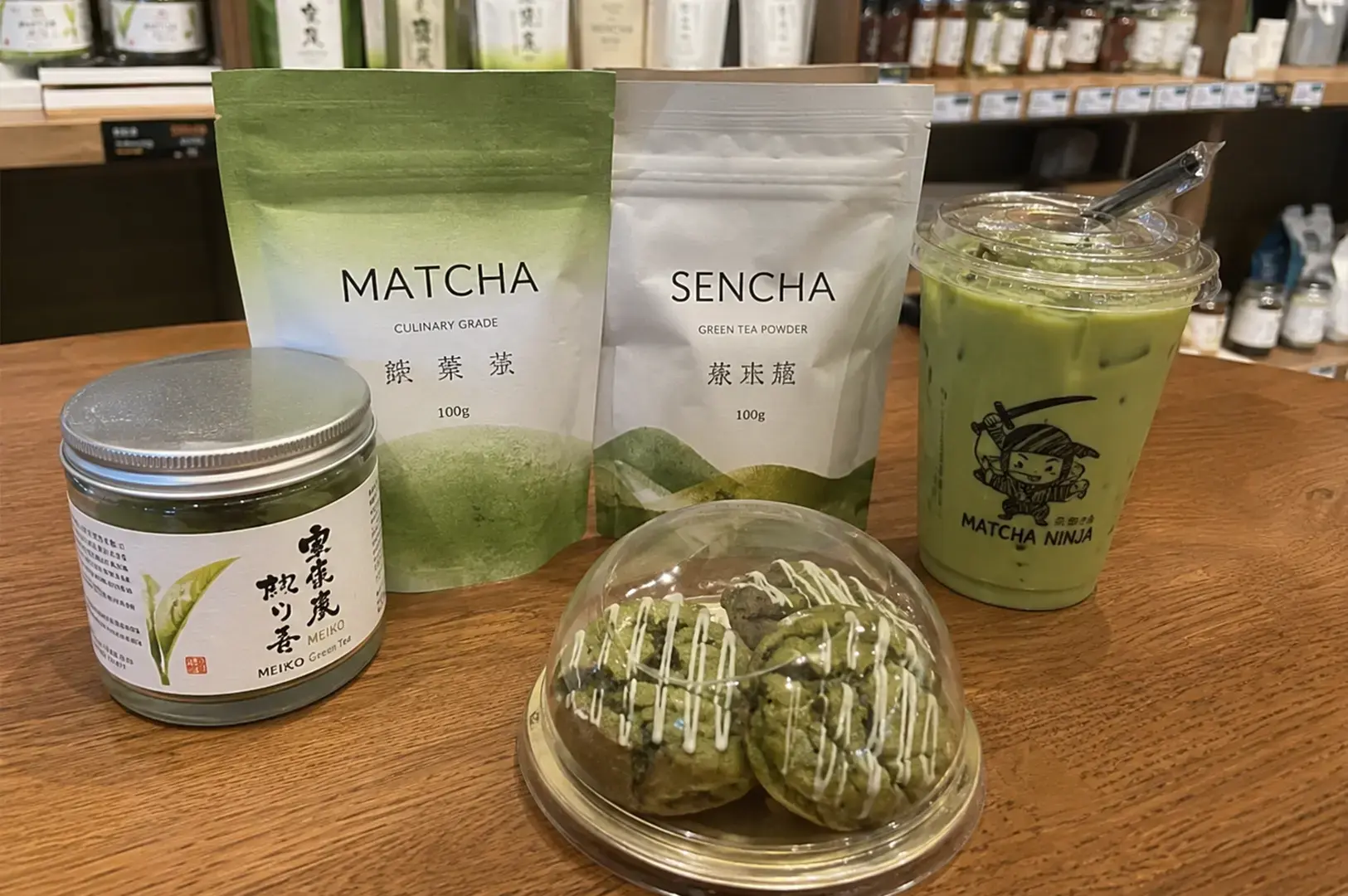 A collection of green tea products is arranged on a wooden surface, featuring a jar of Meiko green tea, bags labeled Matcha and Sencha green tea powder, a plastic cup of iced matcha latte with a ninja-themed logo, and a plastic-domed container holding several matcha-flavored cookies with white icing drizzles.