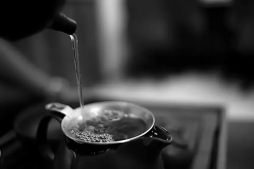 A stream of clear liquid pours from a dark teapot spout into a strainer resting atop another ceramic pot. The black-and-white, close-up photograph captures bubbles forming on the surface of the liquid within the strainer against a softly blurred background.