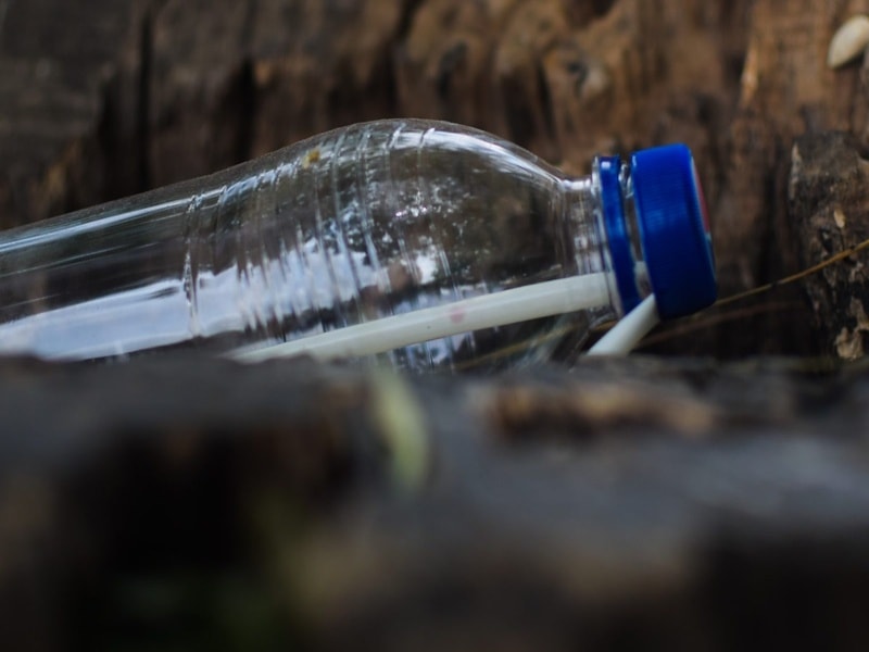 An empty plastic bottle with a blue cap and a plastic straw inside, lying on the ground between pieces of wood.