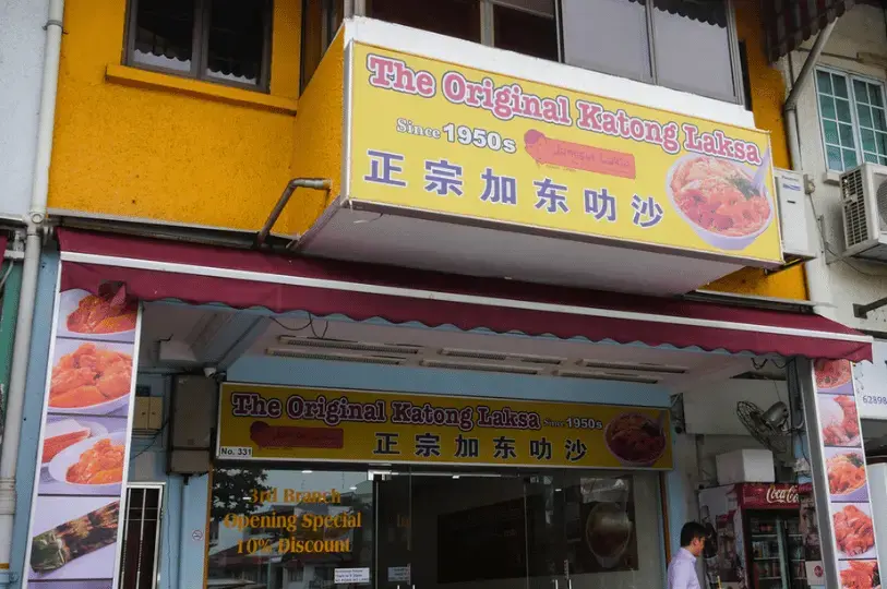 The storefront of "The Original Katong Laksa" (Janggut Laksa) featuring a large yellow banner and awning. The sign notes the brand has been around since the 1950s. A glass entrance shows a clean, modern interior with a "3rd Branch Opening Special" sticker on the window.