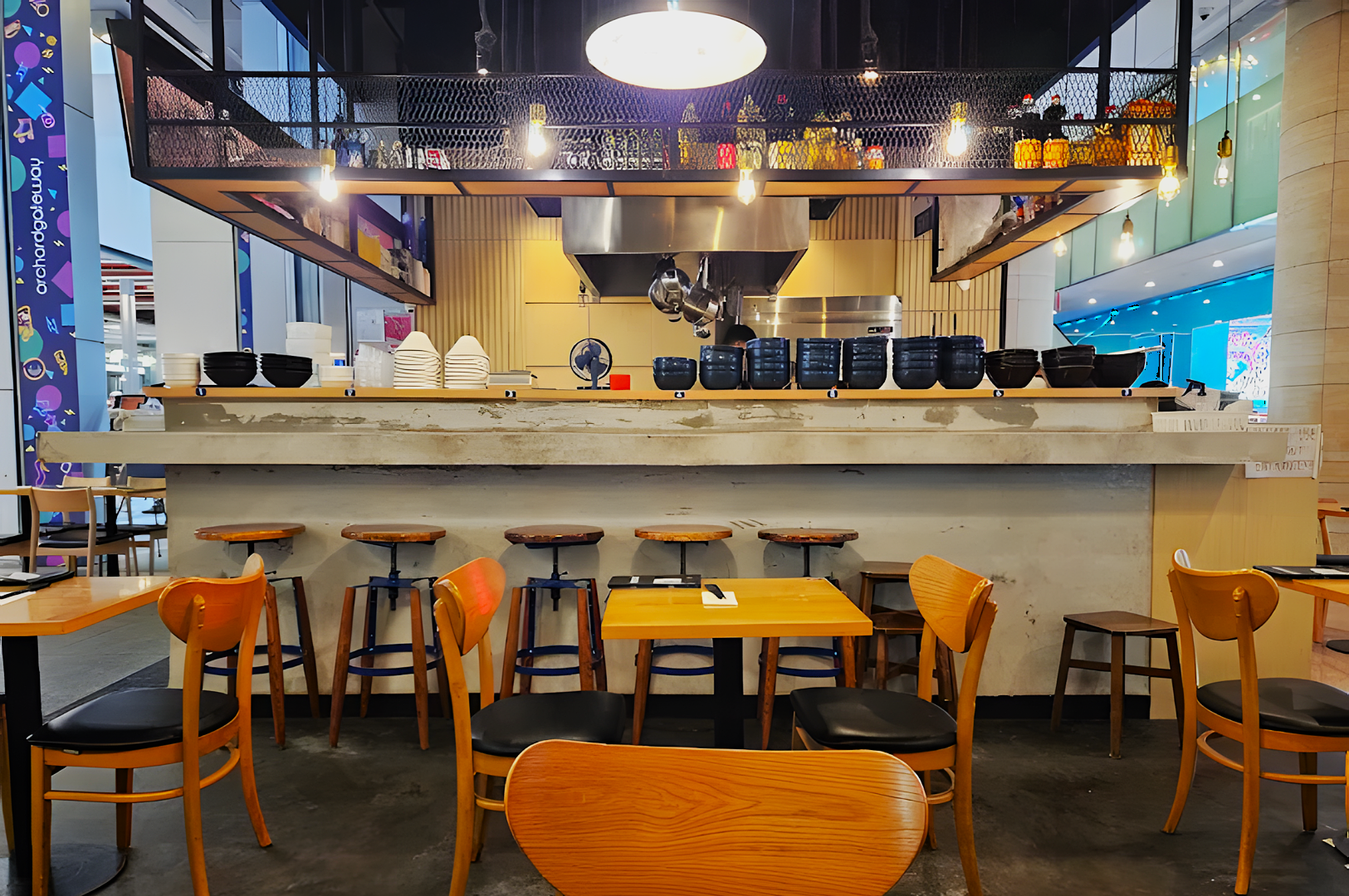 A cozy restaurant with wooden tables and chairs facing a counter lined with empty bowls and plates. Overhead lights create a warm ambiance.
