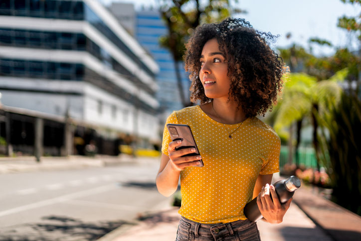 Woman with dark curly hair in a yellow shirt walking down the street holding a cell phone.