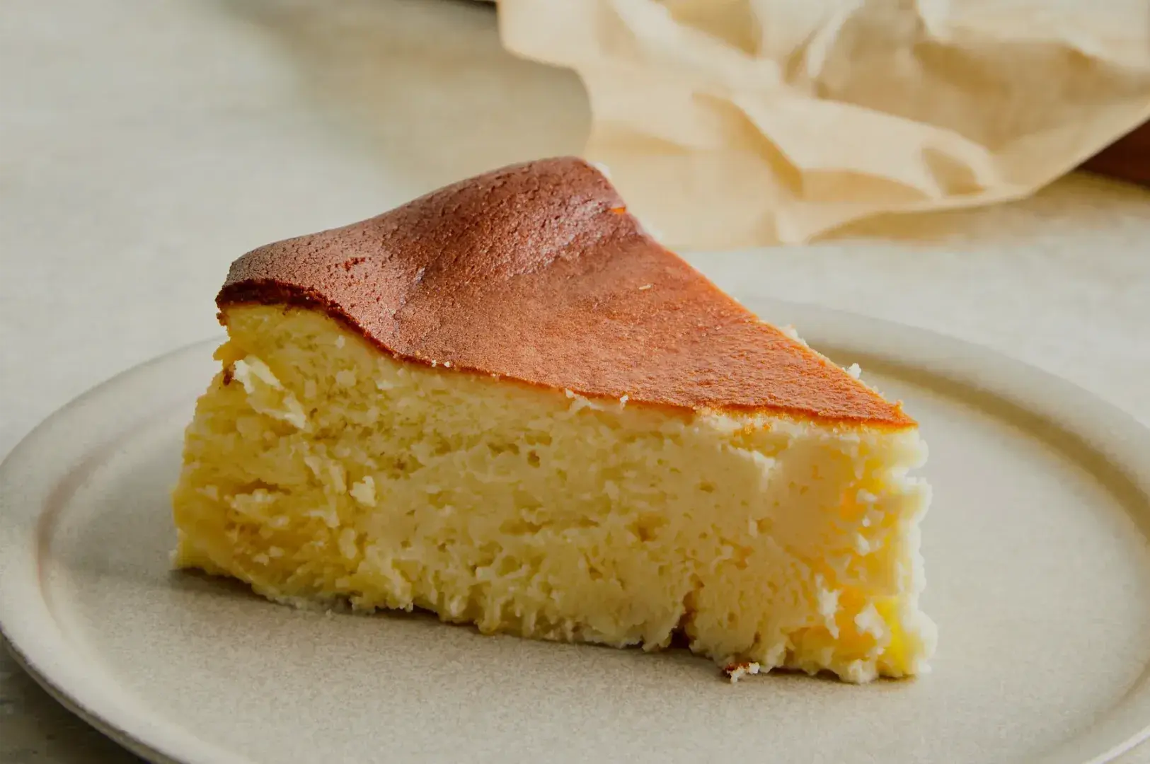 A thick slice of golden Basque cheesecake with a deeply browned top rests on a white plate, creating a warm, inviting dessert scene.