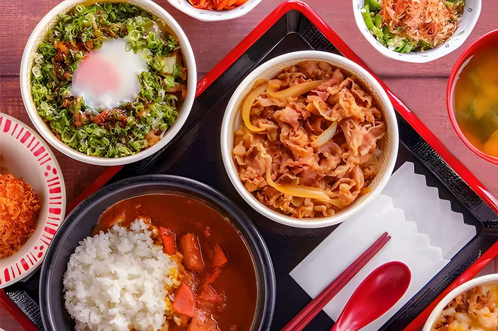 A selection of Japanese food on a marble table including rice, tonkatsu, fried croquettes, a bowl of udon with toppings, and a small bowl of miso soup. Hands hold a bowl of grilled eel over rice.