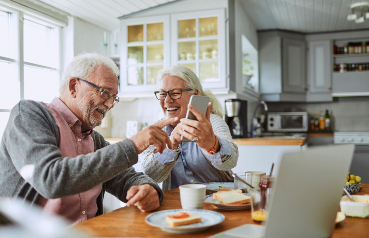 Elderly couple having breakfast and laughing at something on a smartphone. 