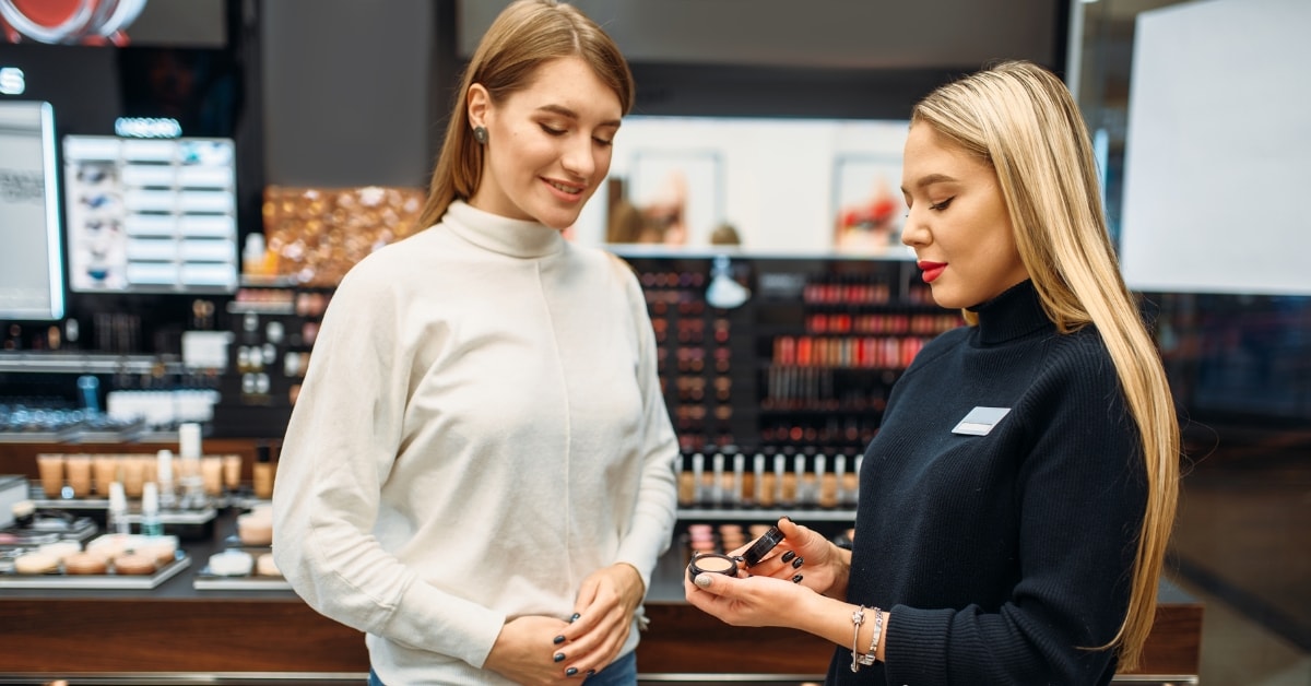 Two women discussing makeup at a store, illustrating examples of Pink Tax in cosmetics pricing.