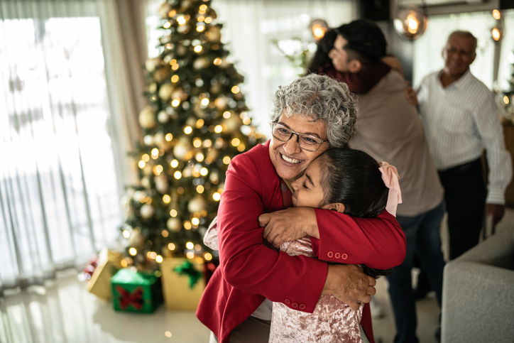 Grey haired grandma hugging her granddaughter by the Christmas tree. 