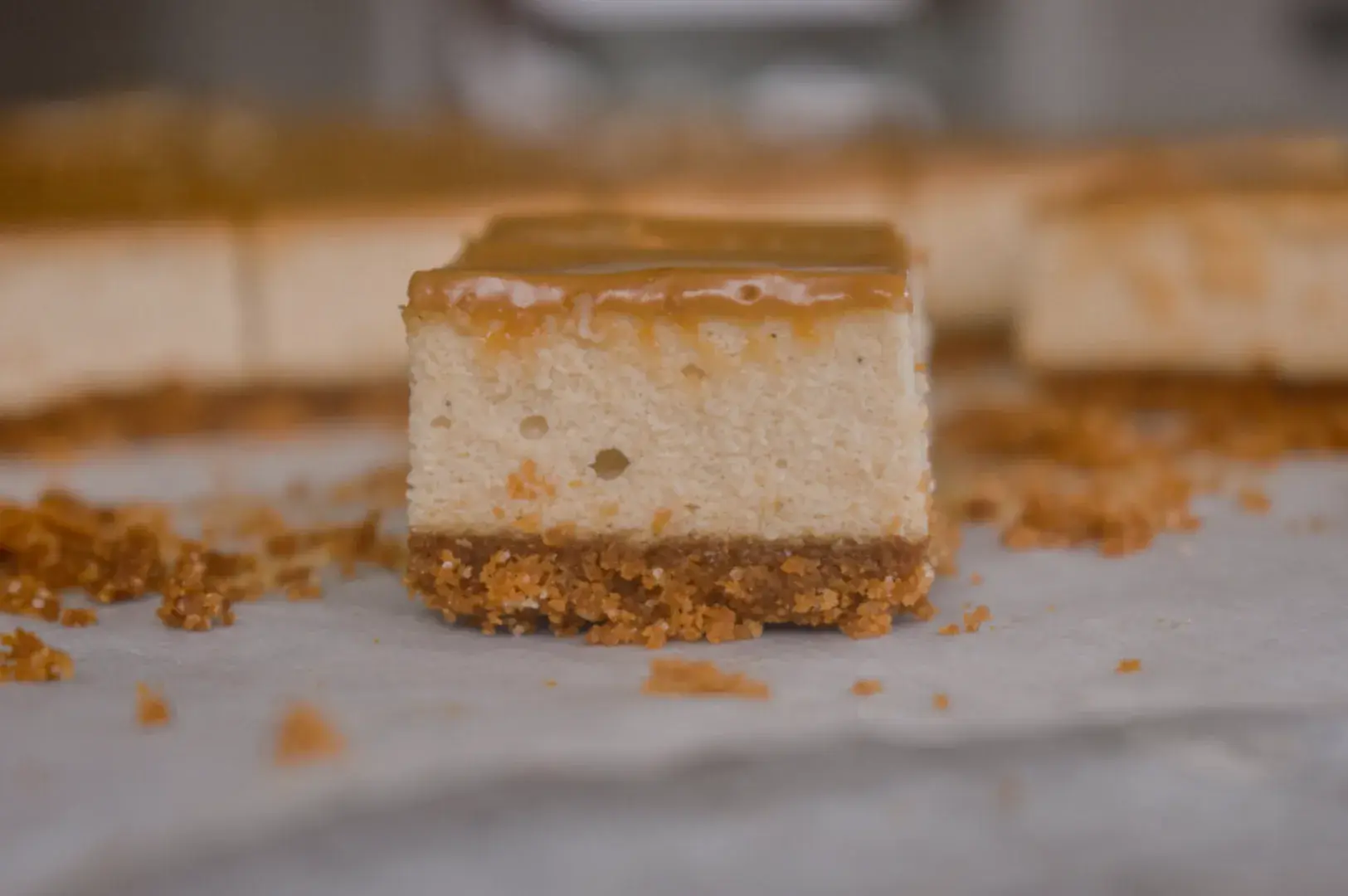 A close-up of a slice of cheesecake with a caramel topping, sitting on a surface scattered with crumbs.