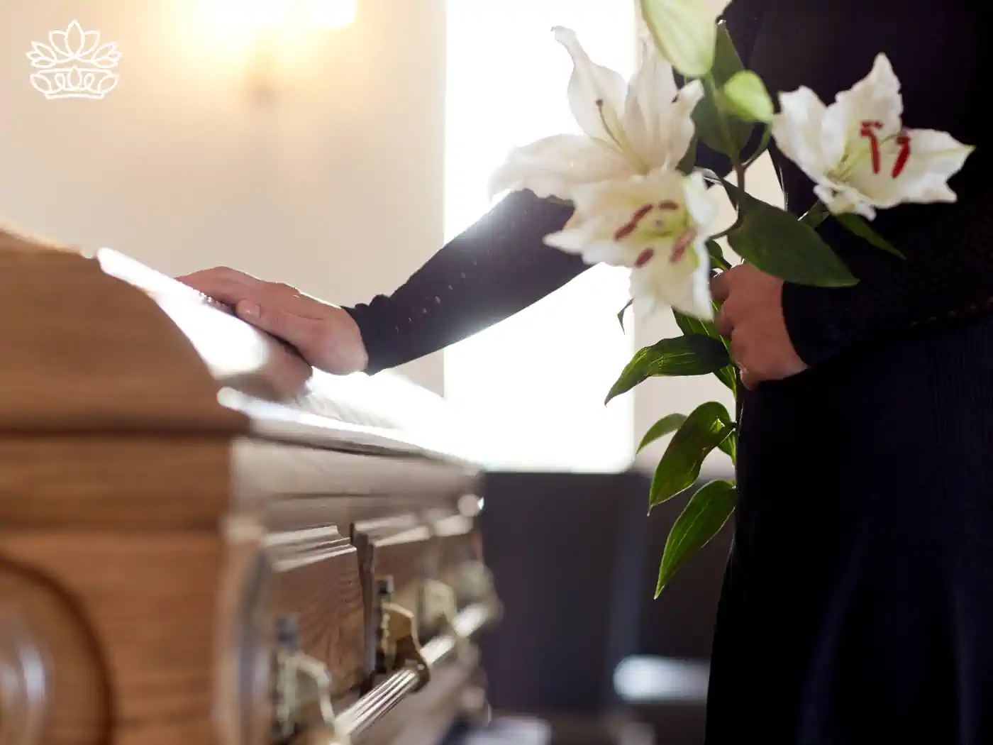 Person placing a hand on a wooden casket with white lilies. Fabulous Flowers and Gifts Funeral Collection.